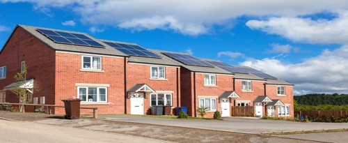 A row of four red brick houses. Each has solar panels on the roof. The sky is blue with fluffy clouds overhead.