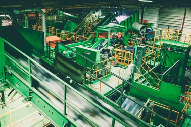 A wide shot of the Re-Gen recycling plant. There are conveyor belts carrying recycling materials around the plant, a person in a high visibility jacket standing at the controls and a waste transfer vehicle emptying recycling onto the sorting line.