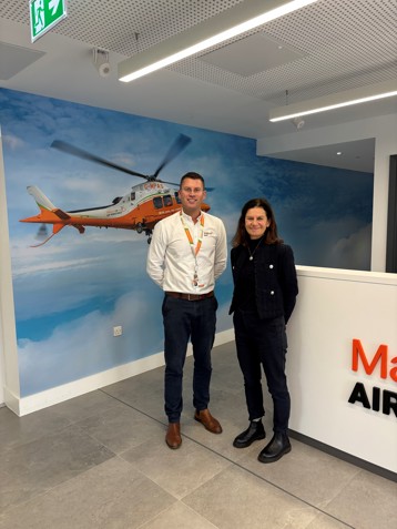 Two individuals standing in a modern office environment in front of a mural featuring an orange and white helicopter in flight, next to a white reception desk.