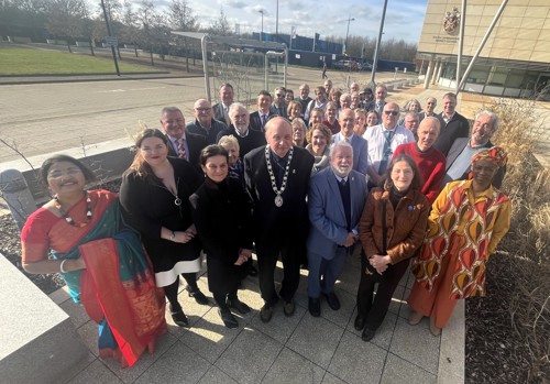 A large group of people standing together outdoors in bright sunlight, arranged in several rows and looking up toward the camera. The group is positioned on a paved area next to landscaped planting and a modern building. Some individuals are wearing formal chains of office, and the group includes people in a wide variety of clothing styles. Trees, parked vehicles and additional buildings are visible in the background.